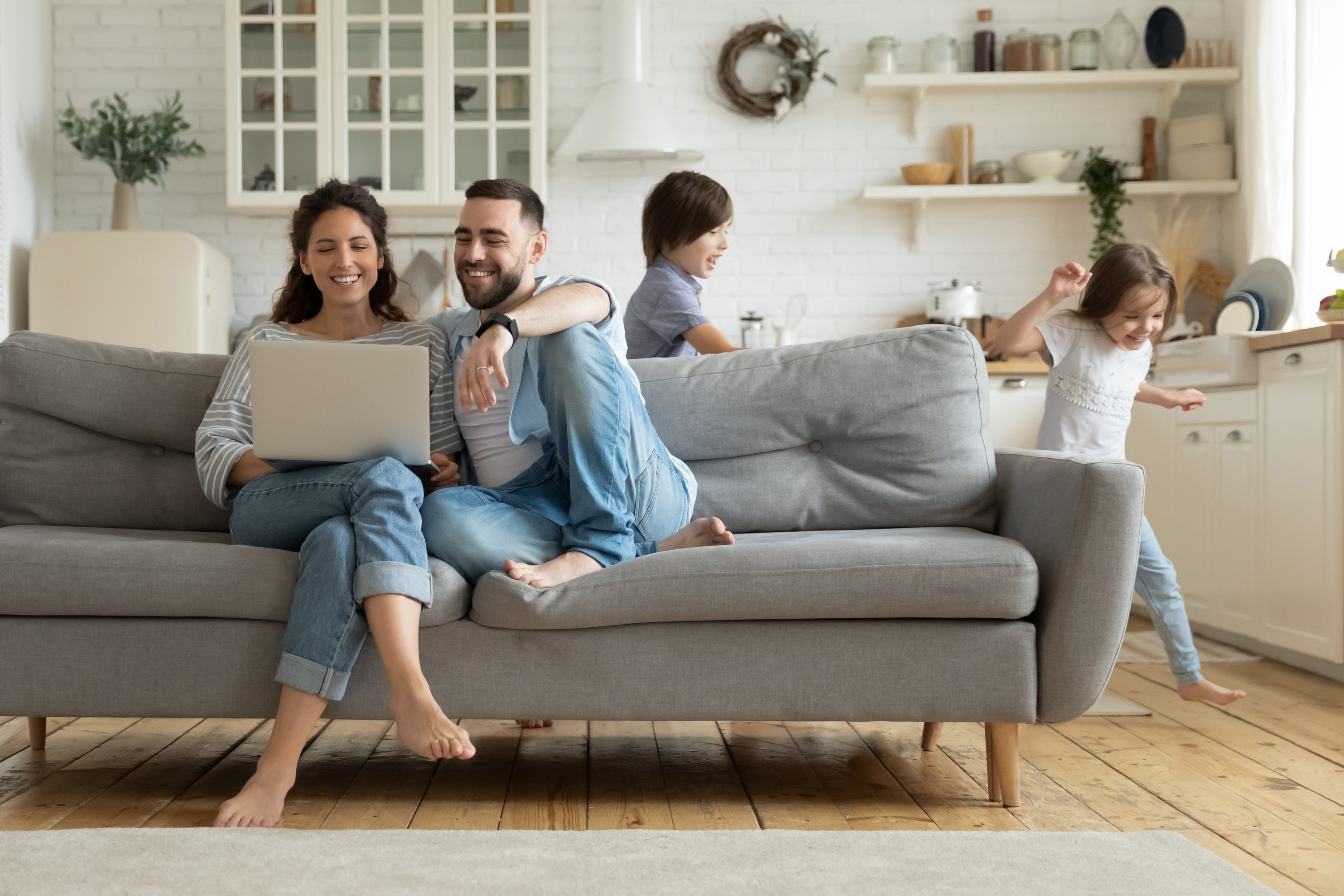 Couple sitting couch looking at a laptop while kids run in the kitchen