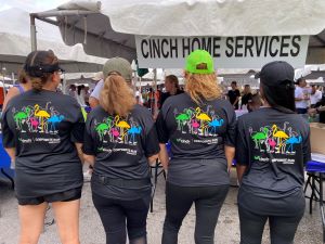 At the 2022 Mercedes-Benz Corporate Run, Cinch employees show off the back of their black race shirts, which showcase the Cinch logo and a flock of colorful flamingos wearing sneakers 