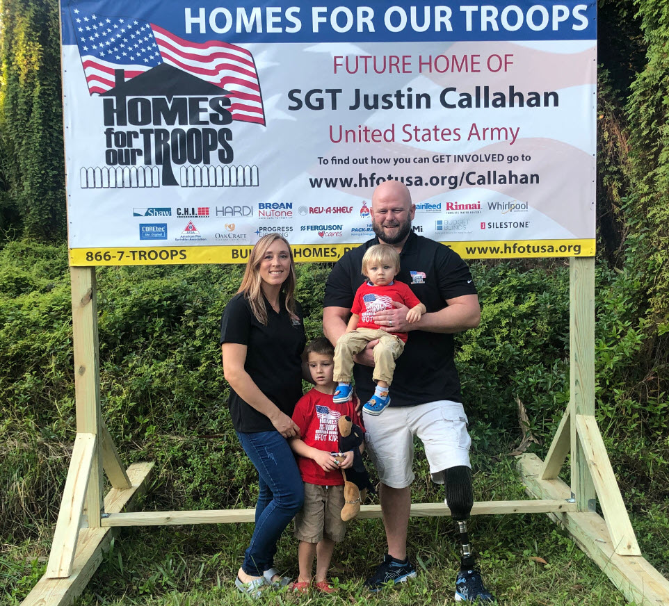 Sergeant Justin Callahan, his wife and two children standing in front of a large Homes for Our Troops display
