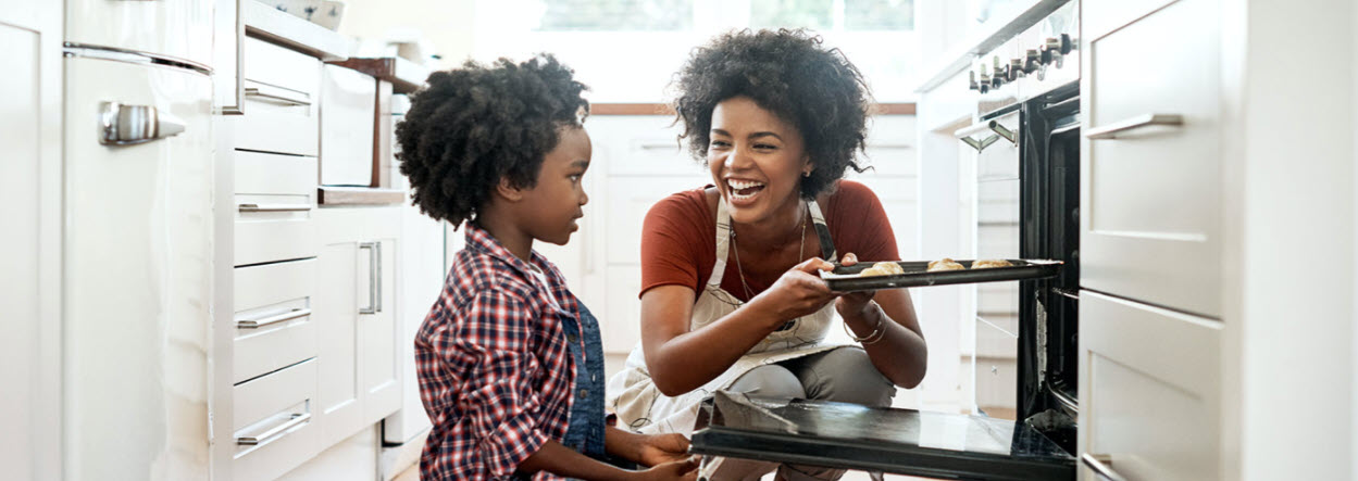 Mother and young child in white kitchen placing a pan of food into the oven.