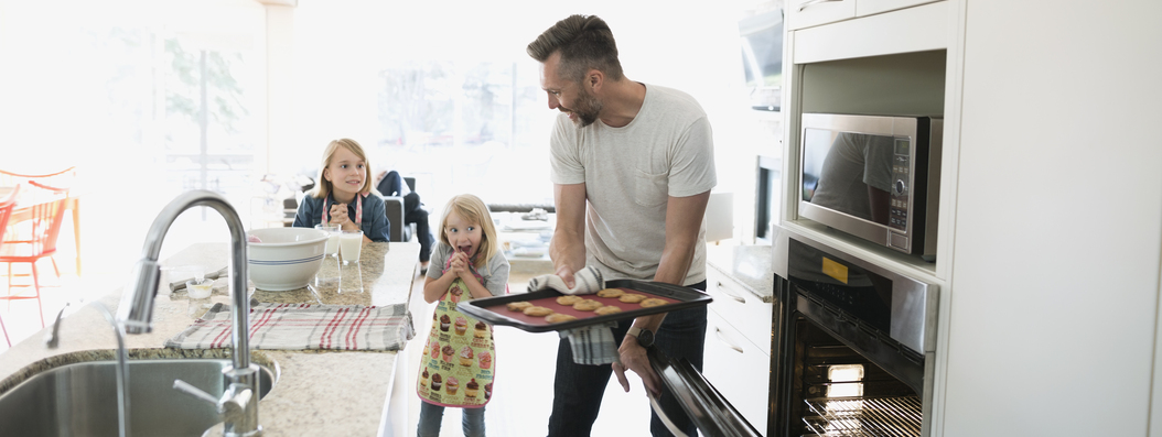 Father pulling out baked cookies out of the oven while his two young daughters look on excitedly