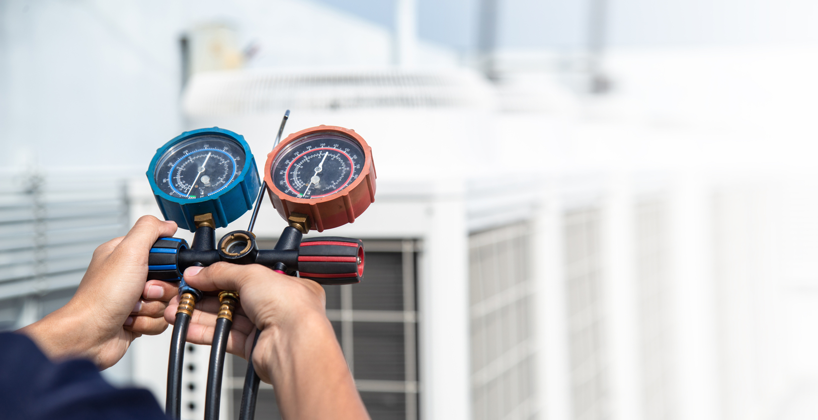 Repairman holding blue and orange measuring HVAC gauges towards an outside HVAC unit
