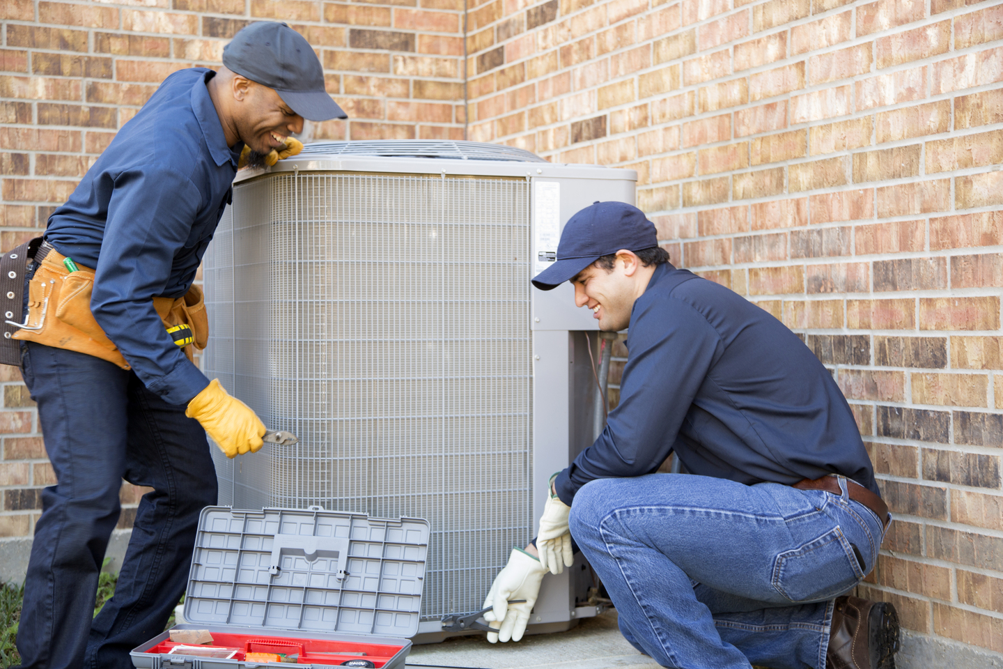 Multiethnic team of repairmen fixing a gray, freestanding air conditioning unit near a brick wall
