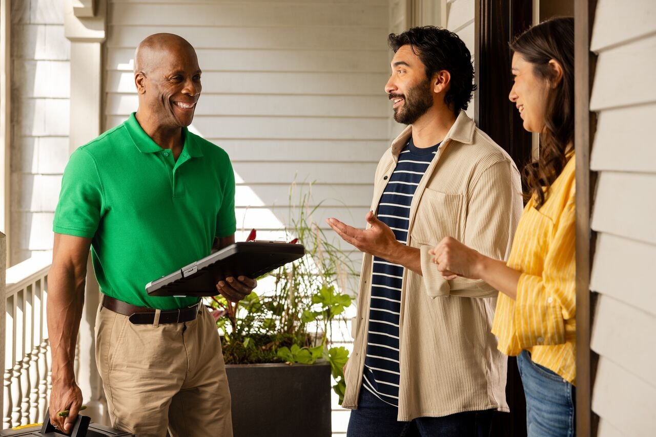 Smiling home warranty technician in green polo shirt holding a tablet, discussing home warranty plans with a homeowner couple at their front door