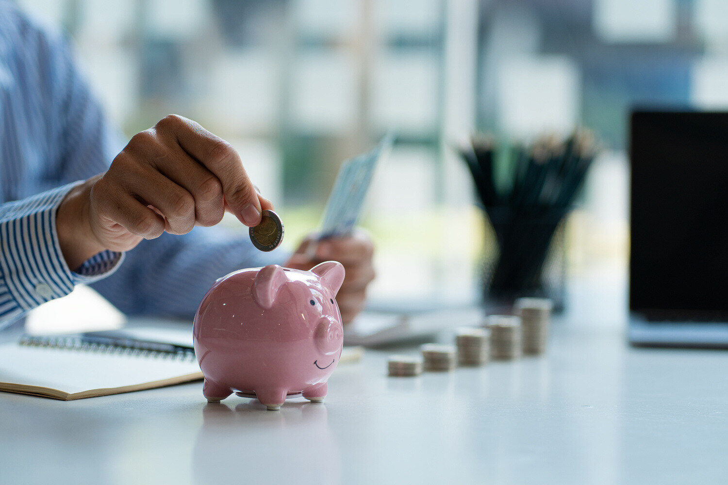 Hands of a businessman dropping coins into a pink piggy bank with coins stacked on the table to save for unforeseen events